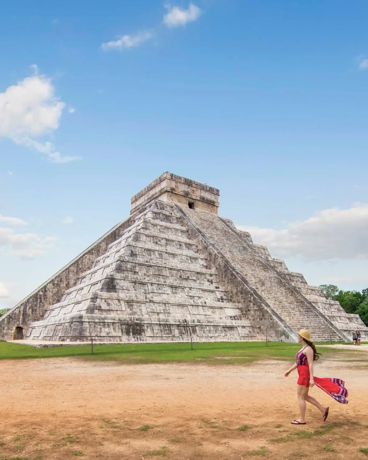 A woman in red dress walking on dirt near a pyramid.